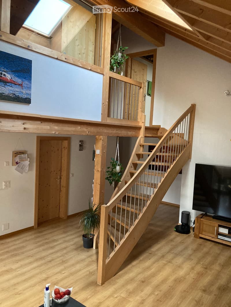 A spacious, well-lit living area with wooden flooring, a staircase leading to an upper level, a television on a wooden stand, and a few decorative plants.