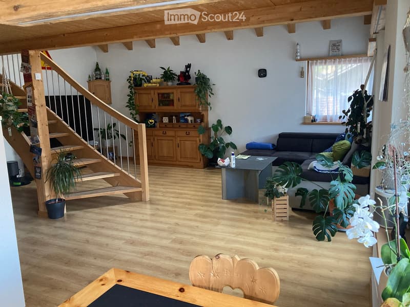 Bright living room with wooden floors, exposed wooden beams, a staircase leading to an upper level, a sofa, coffee table, and a wooden cabinet with glass doors.