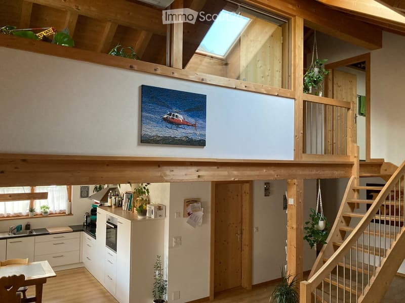 Open-plan living and kitchen area with exposed wooden beams, staircase leading to a loft area, modern kitchen with white cabinets, sink, and oven, and a painting on the wall.