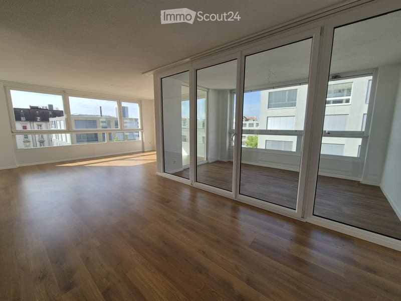 Empty room with wooden flooring and large windows, featuring sliding glass doors leading to a balcony area.