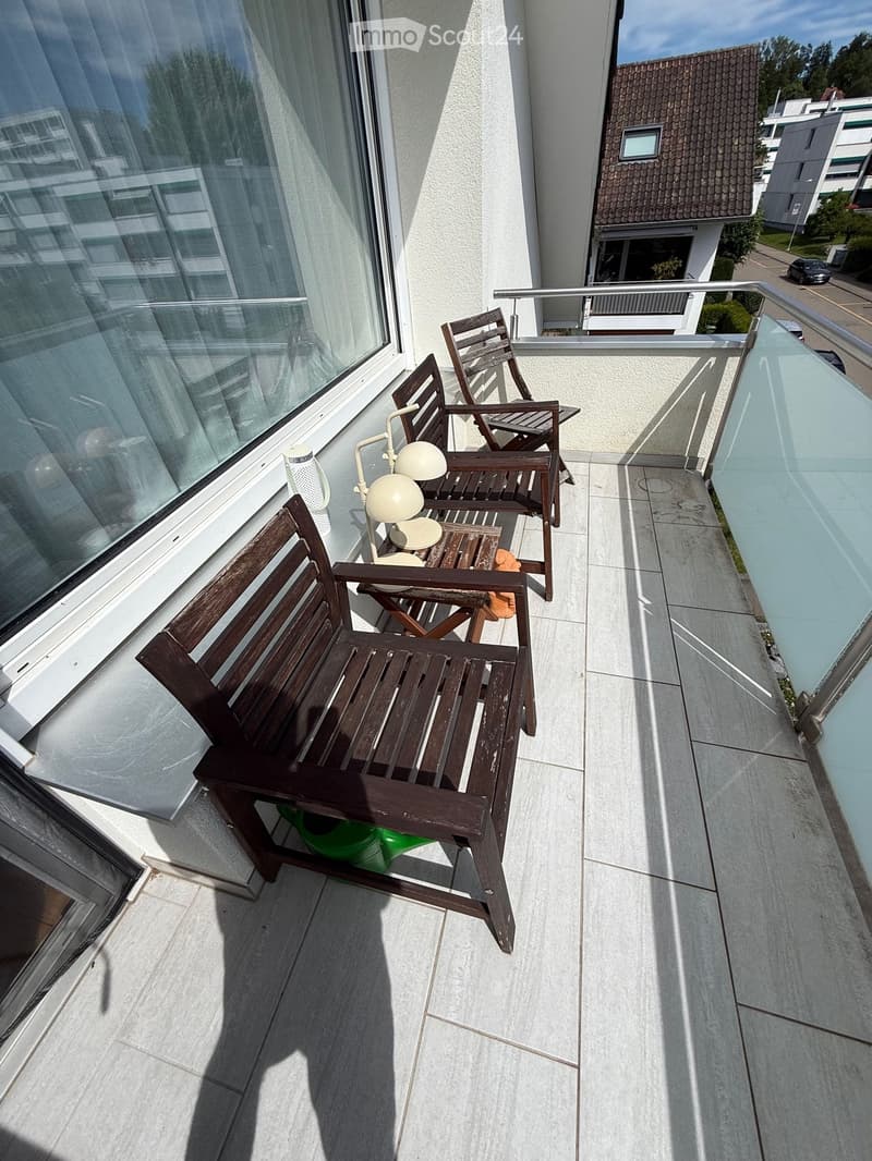 Balcony with wooden bench, chairs, and a table lamp, overlooking a residential area with buildings and trees.