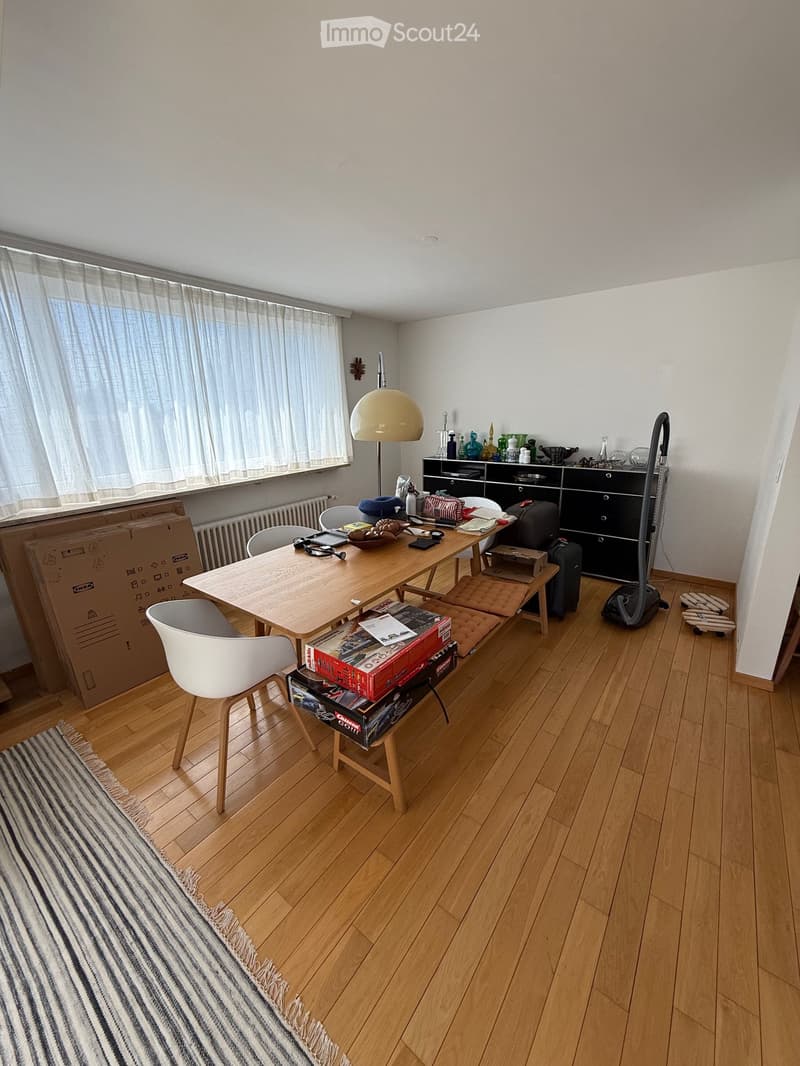 A furnished room with wooden flooring, a large window with curtains, a wooden table with chairs, a lamp, a cabinet, and various items including a vacuum cleaner and suitcases.