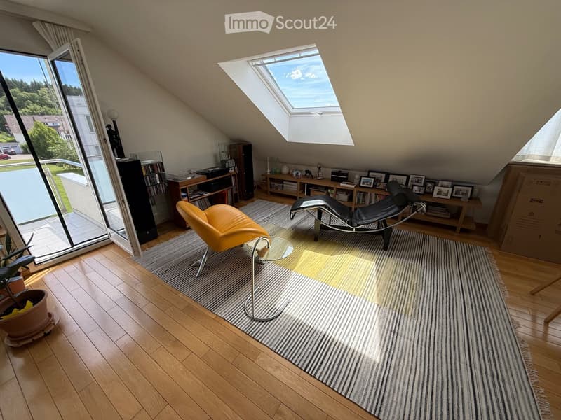 Bright attic room with wooden floors, large skylight, and large window overlooking a garden. Features a yellow chair, a glass coffee table, a black lounge chair, a bookshelf, and a potted plant.