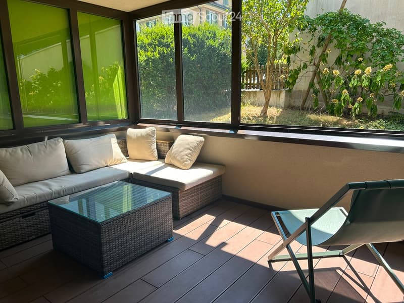 Balcony with a wicker sofa, glass coffee table, and a single chair, overlooking a garden with trees and shrubs.