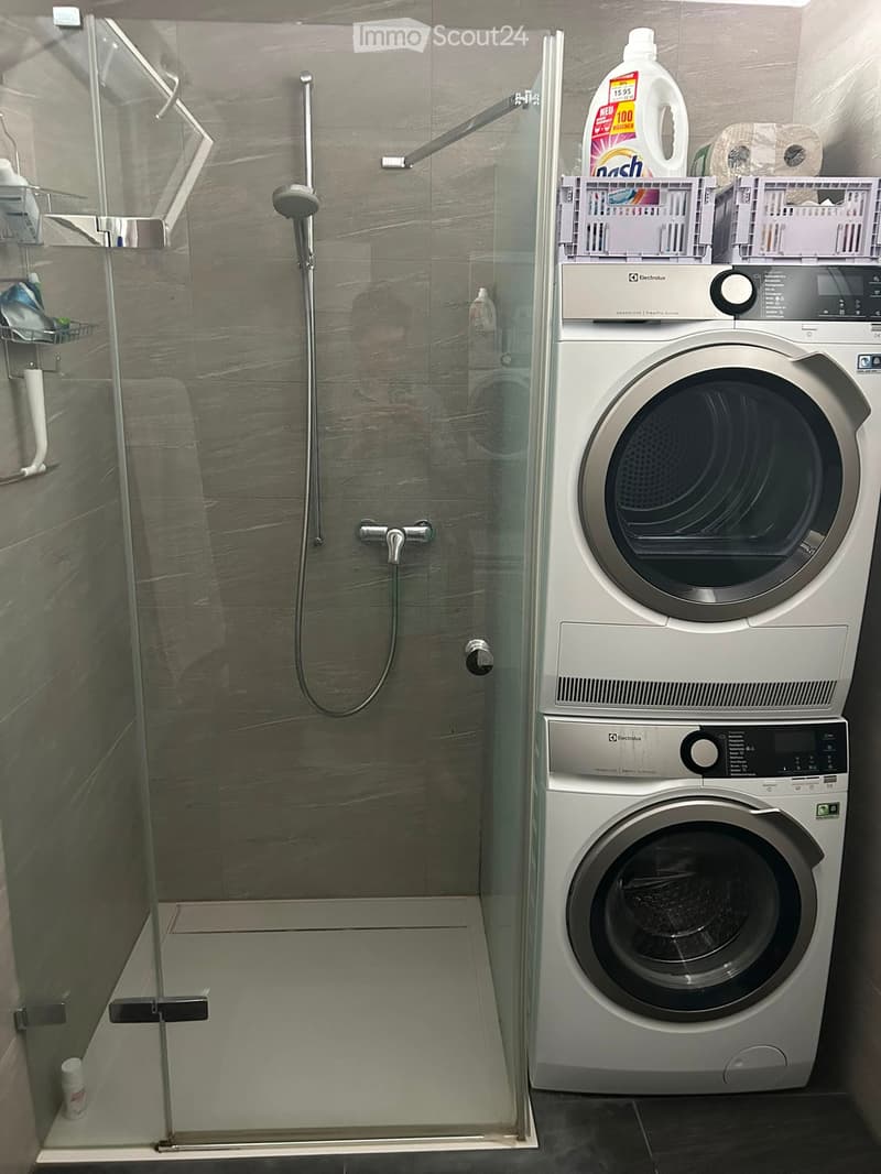 Modern bathroom with a glass-enclosed shower and a stacked washer/dryer unit. Includes a detergent bottle, toilet paper, and a shower shelf with various items.