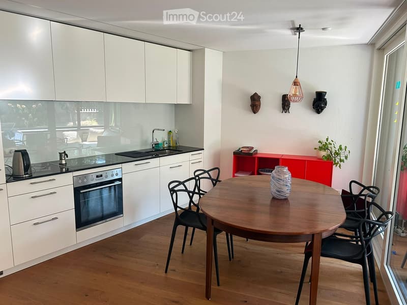 Modern kitchen with white cabinets, a wooden dining table, black chairs, and a red sideboard. The kitchen features an oven, sink, and various appliances.