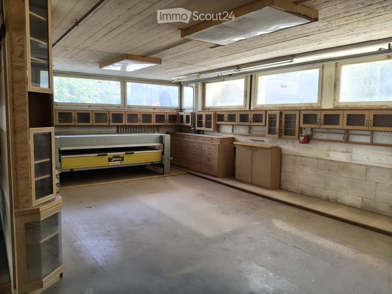 Workshop with wooden cabinets, worktop, and a saw. Exposed wooden ceiling and large windows.