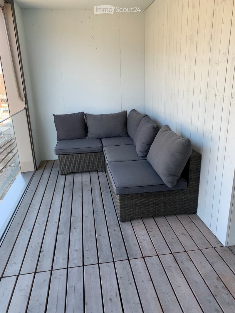 Wooden balcony with a grey wicker sofa and matching cushions, white walls, and a glass railing.