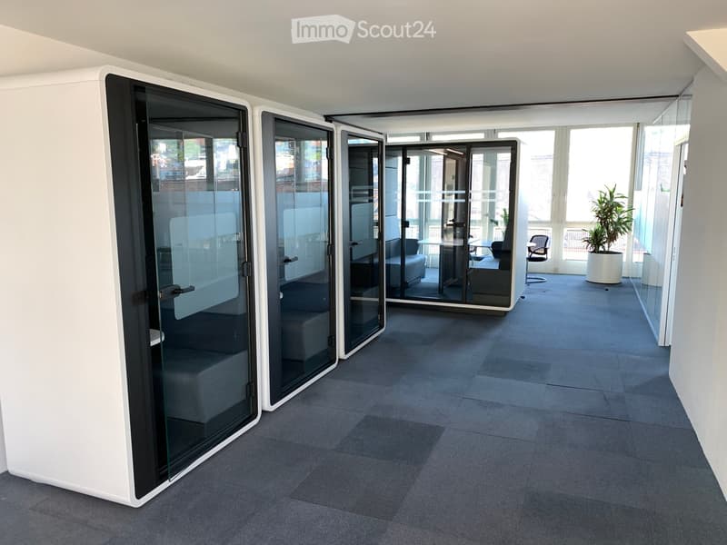 Modern office space with grey carpet, white walls, glass partitions, and a small seating area with a sofa, chair, and table.