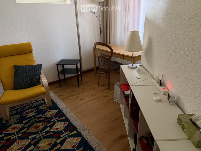 A cozy, well-lit room with a yellow armchair, a small side table, a desk with a chair, a lamp, and a shelf with various items.