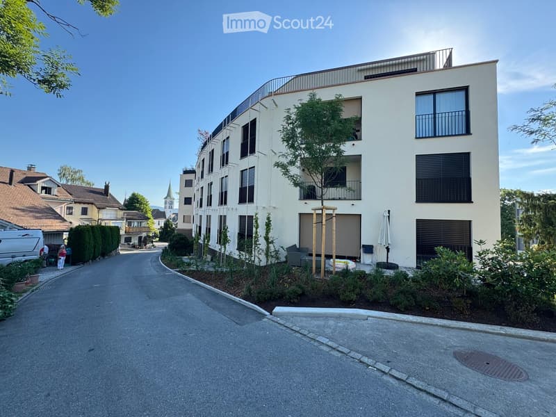 Modern multi-story residential building with balconies, large windows, and surrounding greenery. The building features a sleek, contemporary design with a mix of light-colored walls and dark window frames.