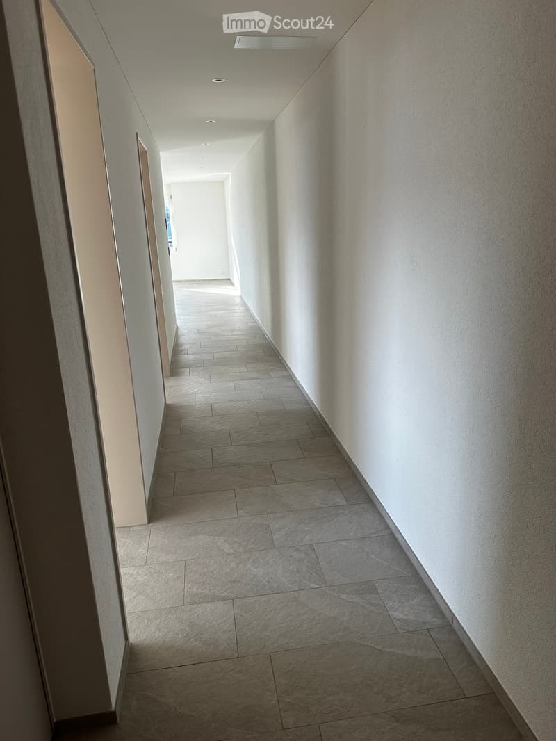 Clean, modern hallway with tiled flooring and white walls, featuring recessed lighting and a visible door at the end.