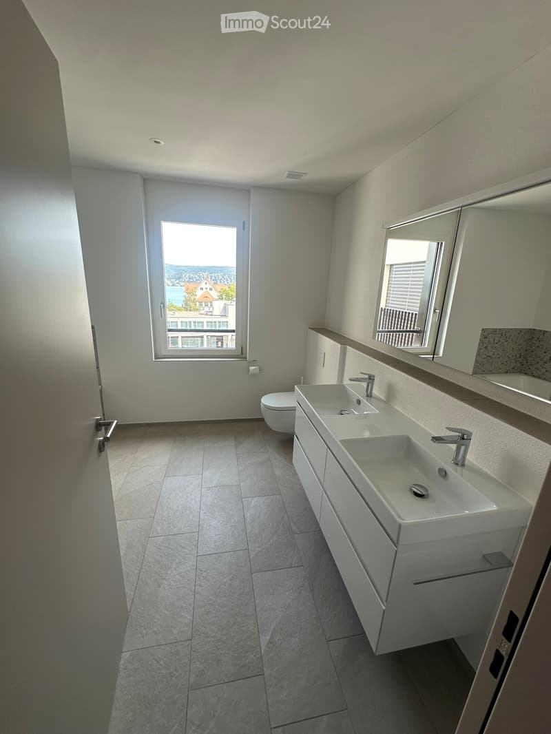Modern bathroom with two sinks, a toilet, a large mirror, and a window offering a scenic view.