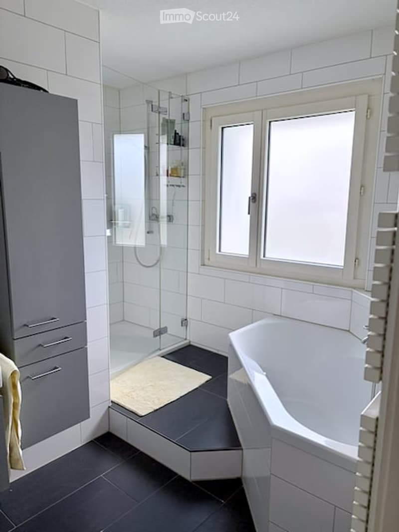 Modern bathroom with white tiles, a bathtub, a glass-enclosed shower, a grey cabinet, and a small rug on the floor.