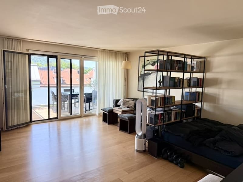 Bright, open living space with wooden flooring, large glass doors leading to a balcony, a sofa, a bookshelf, a fan, and a bed.