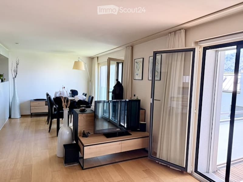 Modern, open-concept living and dining area with hardwood flooring, large windows, and a view. Features a sofa, coffee table, dining table with chairs, a mirror, and decorative vases.