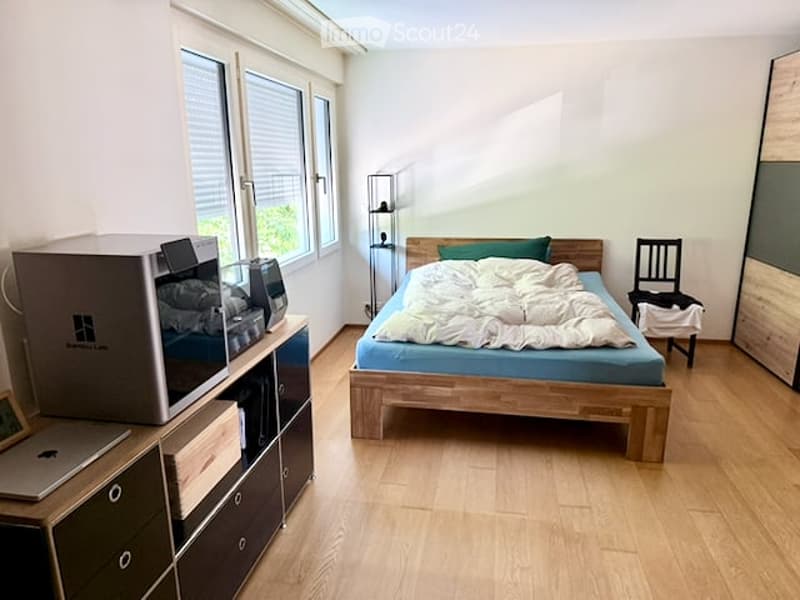 Cozy bedroom with wooden flooring, large windows, a bed with white bedding, a nightstand, a chair, a laptop on a cabinet, and a microwave on a side table.