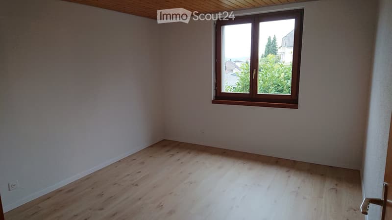 Empty room with light wooden flooring and white walls, featuring a large window with wooden frame and view of greenery outside.