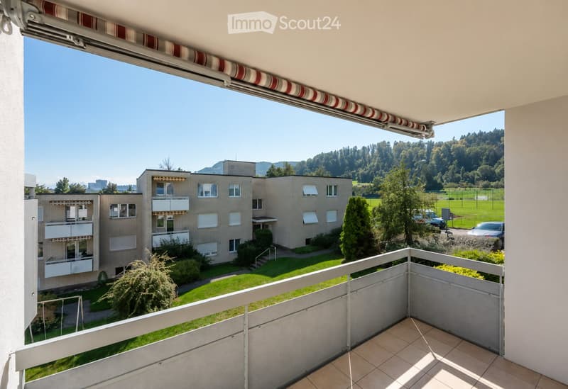 Balcony with tiled floor, white railing, and a view of a multi-story building, green lawn, and surrounding landscape with trees and hills.