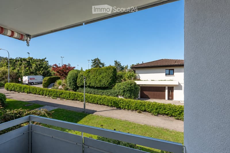 View from a balcony of a two-story house with a garage, surrounded by well-maintained hedges and a driveway.