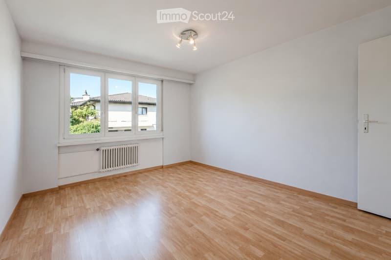 Empty room with light wooden flooring, white walls, large window with view, radiator, and ceiling light fixture.