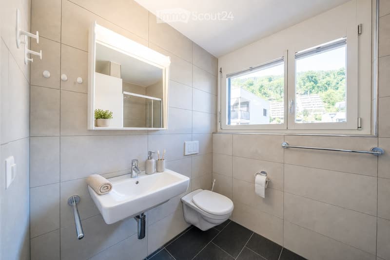 Modern bathroom with a white sink, chrome faucet, toilet, large mirror with built-in lighting, towel, toothbrush holder, soap dispenser, and a small plant on the mirror shelf. The room has beige tiled walls and a dark tiled floor.