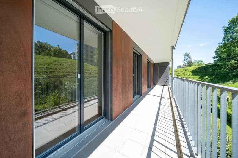 Modern balcony with large glass windows, wooden paneling, metal railing, and a view of a green hillside.