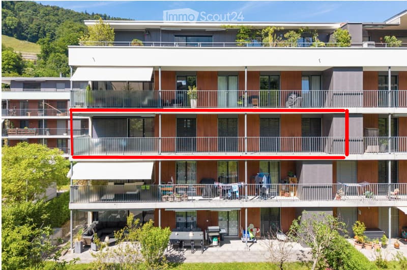 Modern multi-story residential building with balconies, large windows, and contemporary design. Features include wooden paneling, metal railings, and a scenic hillside backdrop.