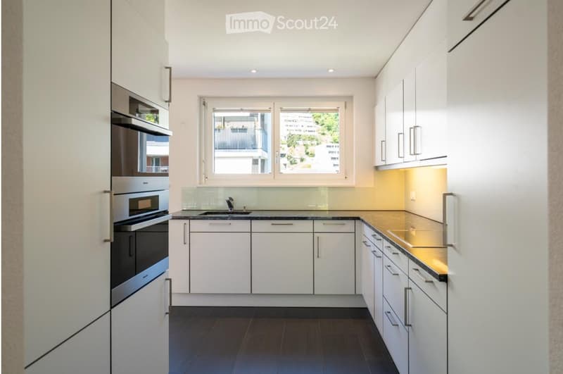 Modern kitchen with white cabinets, stainless steel oven, sink, and a window overlooking a balcony.