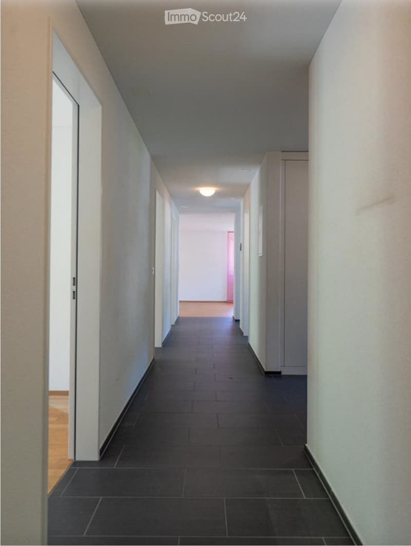 A long, narrow hallway with dark tiled flooring and light-colored walls. The corridor features several doors on both sides and a ceiling light fixture.