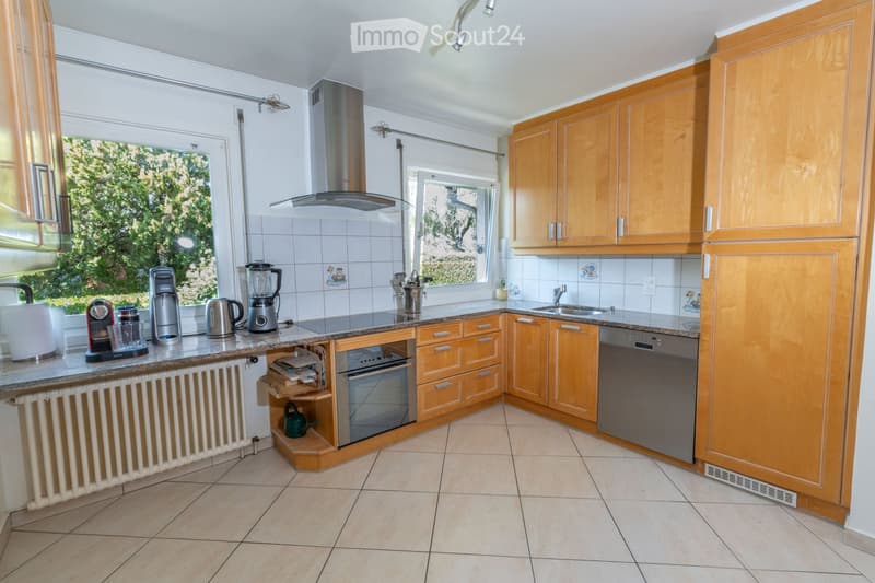 Bright kitchen with wooden cabinets, granite countertop, stainless steel appliances including a coffee maker, blender, oven, and dishwasher. Natural light from windows, tiled backsplash, and tiled floor.