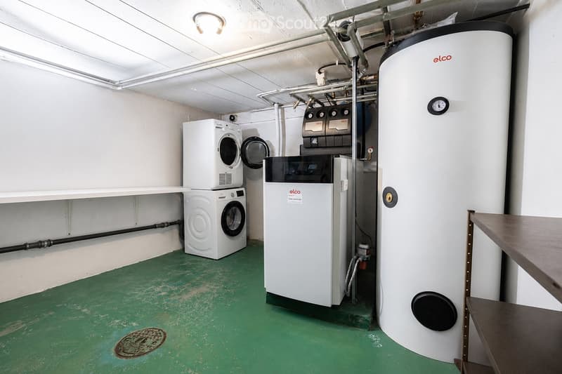 Utility room with green flooring, white washing machine, dishwasher, and water heater, with exposed pipes and a small table.
