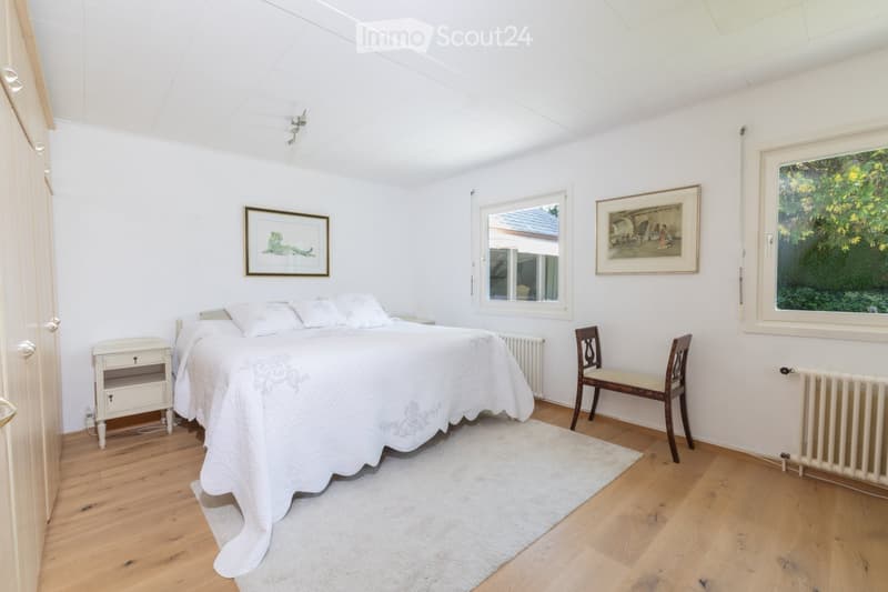 Bright bedroom with wooden flooring, white walls, and a large window offering a view of greenery. Features a neatly made bed with white bedding, a nightstand, a wooden chair, and two framed pictures on the walls.