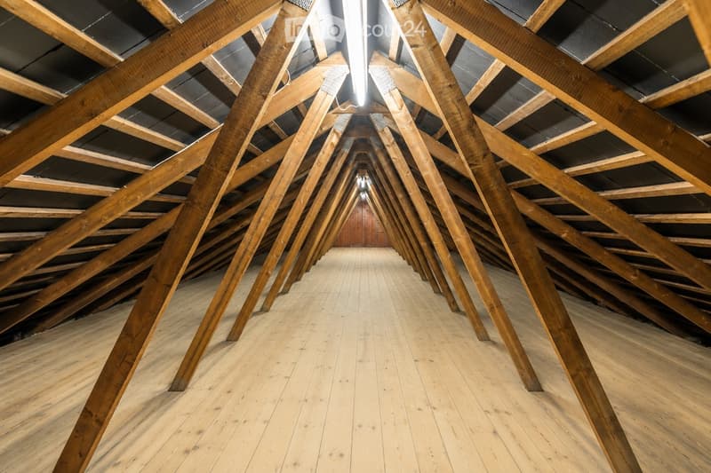 An empty attic space with exposed wooden beams and a wooden floor, illuminated by a single overhead light fixture.
