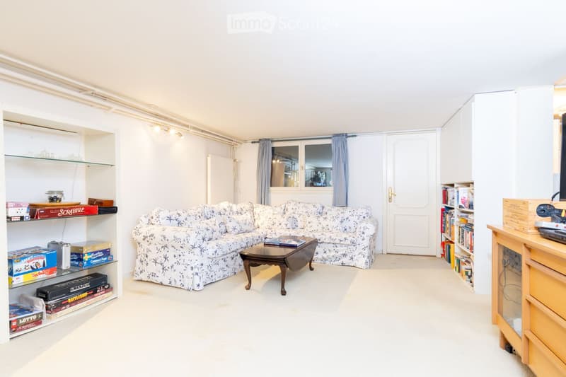 A cozy living room with a patterned sofa, a wooden coffee table, built-in shelves filled with books and games, and a large window with blue curtains.