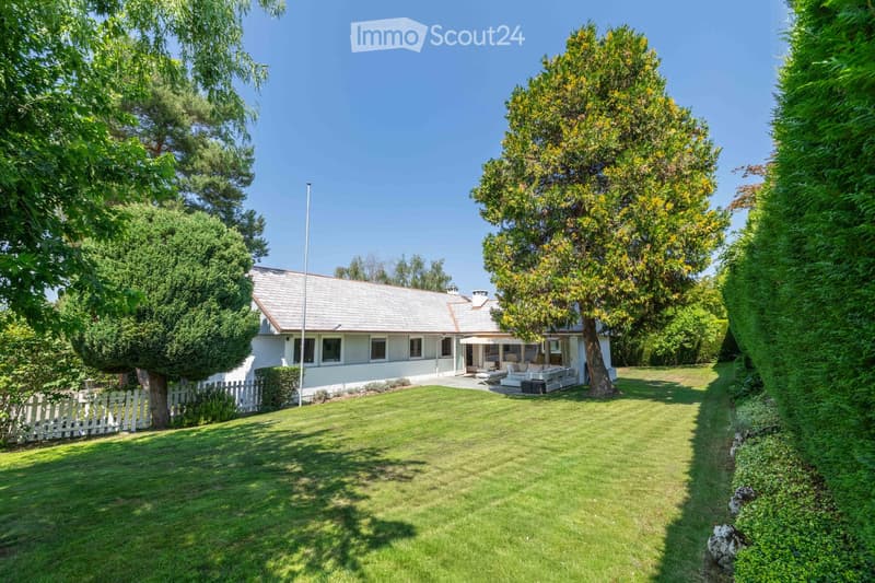 A well-maintained garden with a white picket fence, a single-story house with a grey roof, and outdoor seating area with a sofa and chairs.