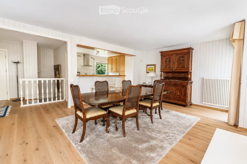 Dining room with wooden dining table, six chairs, a large wooden cabinet, a lamp, and a picture frame on the wall. The room features light wooden flooring and a soft, light-colored rug.