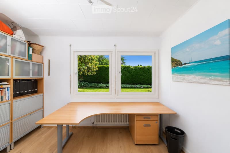 A well-lit room with a large wooden desk, a cabinet with books and filing cabinets, and a large picture frame on the wall. The room has wooden flooring and large windows with a view of a green garden.