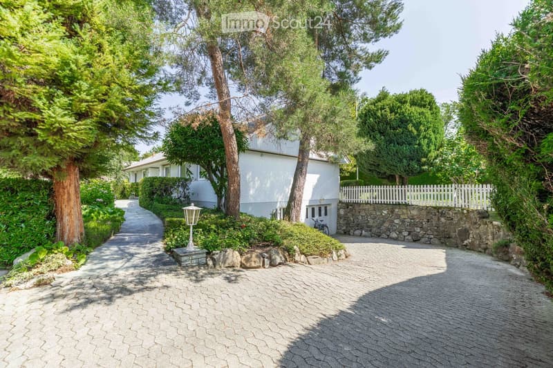 A well-maintained garden path leads to a white house surrounded by lush greenery, trees, and a stone wall with a white picket fence.