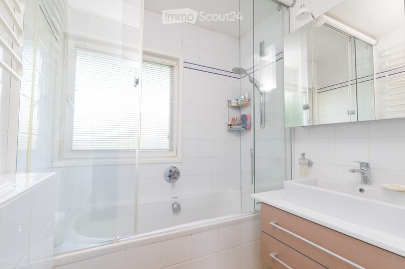 Modern bathroom with a bathtub, glass-enclosed shower, sink with wooden cabinet, and a mirror above the sink. The walls are tiled with white tiles and blue accents.