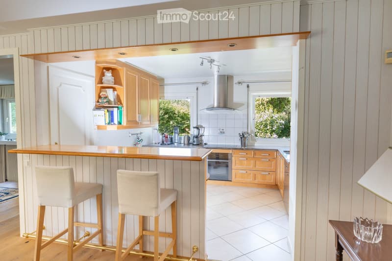 Bright kitchen with wooden cabinets, a bar counter with two stools, and a modern oven. The space features a tiled floor and ample natural light from large windows.