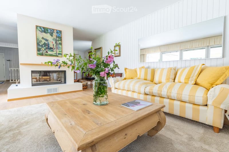 Bright living room with a yellow and white striped sofa, wooden coffee table, vase with flowers, fireplace, and large mirror. Light wood flooring and white walls.