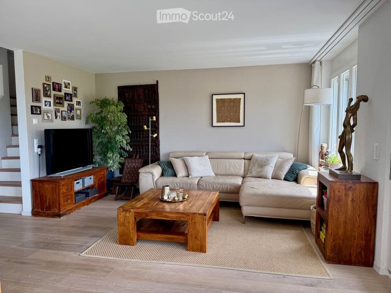 Modern living room with a beige sectional sofa, wooden coffee table, flat-screen TV on a wooden cabinet, floor lamp, decorative picture frame, plant, and a small statue on a side table.