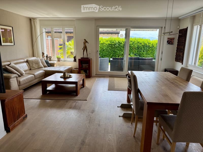 Open-plan living and dining area with light wood flooring, large windows, and a sliding glass door leading to a patio. Features a cream leather sofa, wooden coffee table, dining table with chairs, and decorative items.