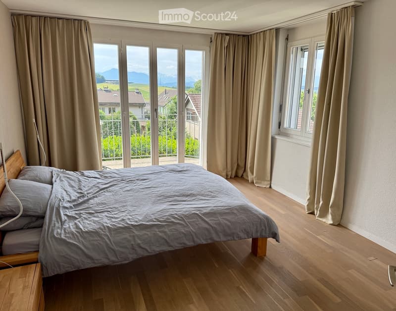Bright bedroom with wooden flooring, large window and sliding glass doors leading to a balcony, offering scenic mountain views. Features a neatly made bed with grey bedding and a wooden nightstand.
