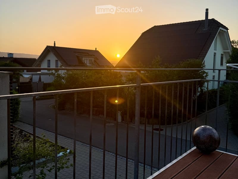 Balcony with metal railing, wooden table, and decorative sphere, overlooking a residential street at sunset.