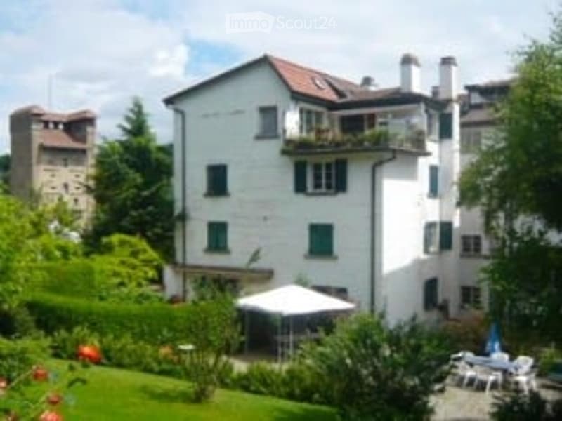 A multi-story white building with green shutters, featuring a balcony and a well-maintained garden with outdoor furniture.