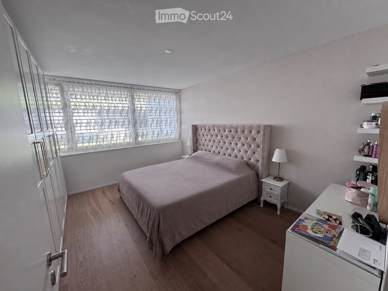 A modern bedroom with a tufted headboard, wooden flooring, and large window with blinds. Includes a nightstand with a lamp, a small table with various items, and shelves with personal items.