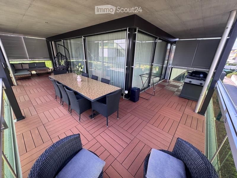 Balcony with wooden decking, glass walls, a dining table with chairs, a sofa, a hanging wicker chair, and a grill.