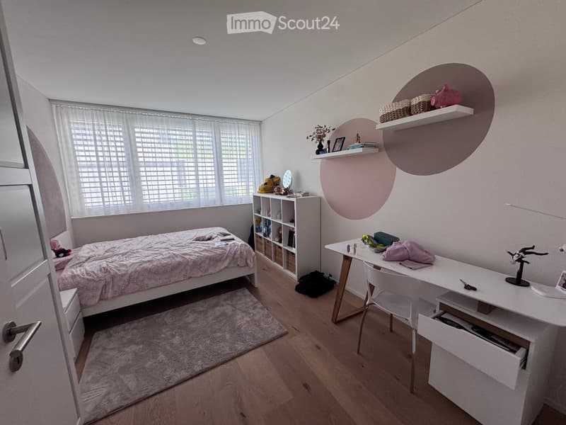 Cozy bedroom with a bed, desk, chair, shelves, and a mirror. The room features wooden flooring and large windows with blinds.